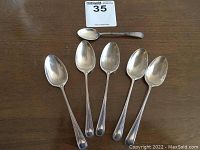 Six spoons laid out on a wooden surface: five matching coffee spoons and one smaller spoon.