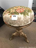 View of the swivel stool showing the padded seat with floral upholstery and gold painted pedestal base with ornate metal legs.