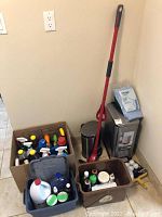Photo showing stainless steel trash can with foot pedal, red handle mop, broom, dustpan, and multiple boxes and tubs holding various cleaning products.