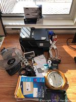Image showing HP Officejet Pro 8600 printer with accessories including office paper, wall clock, small metal fan, digital alarm clock, calculator, bike lock, and various office supplies on floor near window