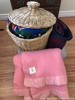 Wicker basket with lid, partially filled with two blankets inside. Pink Eaton wool blanket folded outside basket.
