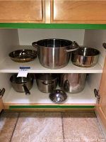 Photo of a shelf with 7 assorted stainless steel pots and pans including large stock pot, smaller pots and a strainer