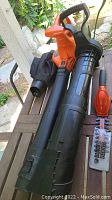 Full view of electric leaf blower with black collection bag, black nozzle, and orange housing, alongside orange battery trimmer part and blade guard.