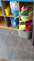 Photo showing various cleaning liquids, sponges, brushes and storage container on floor under a blue shelf.