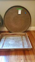 Copper round tray with Indian inspired designs leaning against wall, with rectangular wooden tray with metallic inlay in front on wooden floor.