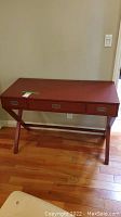 Front view of reddish wooden desk with three drawers and metal handles on an X-shaped leg frame on hardwood floor against wall.