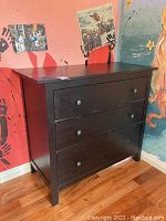 Full view of black wooden dresser with three drawers, showing intact knobs except missing one on top drawer, and some visible surface scratches.