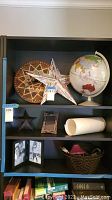 Wide view of the shelving unit showing globe, large wooden clock, metal stars, picture frames, woven basket with ceramic item, and rolled white item.