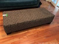 Long rectangular upholstered ottoman on wooden floor with dark legs and patterned fabric.