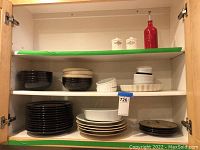 Ceramic plates and bowls organized on three shelves inside a wooden cabinet, showing various colors and sizes including black and cream pieces