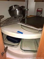 Photo showing stacked metal mixing bowls, metal toast rack, sheet pans on top of a white circular shelf and Pyrex glass baking dishes on the lower shelf.