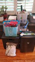 View of entire lot showing shredder, rolling crate, tote with folders, gift bags, and white rolled item in plastic bag on a wooden floor near window