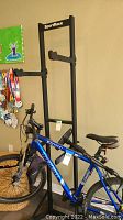 Black metal SportRack bicycle storage rack holding a blue bicycle frame with adjustable arms, placed indoors near a beige wall.