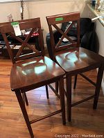 Two wooden bar stools with X-shaped backrests sitting on a wooden floor.