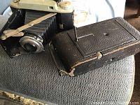 Two vintage folding cameras placed on fabric surface: Kodak Tourist II and Rexo.