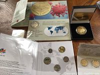 Two COVID-19 commemorative coins in display card and presentation box alongside various coin sets spread on wooden surface