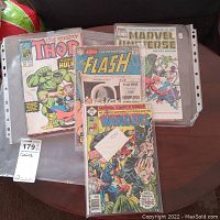 Photo showing a stack of comic books including five Thor comics, Invaders comics issues 18, 34, 35, and 36, The Flash comic, Marvel Universe, Hawkeye, Defenders, and Marvel Saga comics in plastic sleeves.