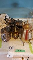 Photo showing six silver plated serving pieces including coffee pot, teapot, coffee urn, sugar bowl, creamer, and small bowl, arranged together on a counter with a note indicating measurements (2.5 to 10 inches tall) and lot number.