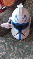Front view of white Clone Trooper helmet with blue markings and black vent details on fabric sofa.