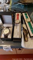 Four watches displayed in their boxes and on a cloth, showing variety of bands and colors including red and green leather bands and metal bands.