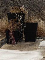Three black woven rectangular planter pots with dried plants and soil, placed outdoors by a poolside on concrete and rocks.