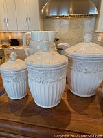 Three white ceramic canisters and two white ceramic pitchers on wood counter in kitchen.