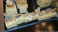 Photo showing eight Cherished Teddies figurines on their original boxes partially wrapped in plastic, arranged in a row.