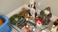 Photo showing two Santa Claus figurines in red and green cloaks, a snowman ornament, a small gift box, and pinecone decorations arranged on the floor near a wall.