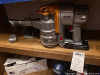 Front view of Dyson cordless stick vacuum on wooden shelf with battery installed; charging dock and power adapter at left; Rowenta box beneath