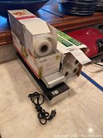 Full view of the vacuum sealer with plastic sealing rolls on top, showing power cord and packaging.