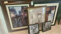 Five pieces of framed wall art leaning against a wall, showing a rainy city street scene with umbrellas, a gold framed art with historical building and musical notes, and smaller black framed black and white nature prints of zebra and elephant.