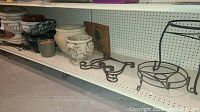 Wide view of the shelf showing several ceramic pots and metal pot stands arranged on a white pegboard shelf in basement area.
