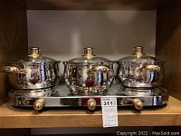 Front view of the entire hot plate with three pots, all featuring gold-colored handles and glass lids, on a wooden surface.