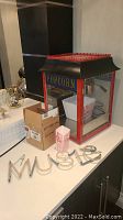Red and black tabletop popcorn popper with glass sides and blue 'POPCORN' sign on front, popcorn boxes in cardboard box next to it.