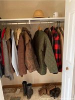Photo showing variety of coats including quilted green, plaid, leather, fur-collared, and white vest hanging in closet, along with pith helmet on shelf and boots on floor.