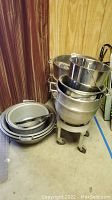 Photo showing stacked commercial metal mixing bowls and pots on a wheeled metal stand against a curtained background.
