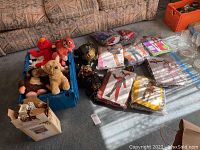 Overall lot contents with blue crate filled with various stuffed animals, several packaged costumes on floor, cardboard box with dollhouse furniture