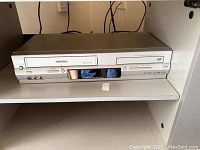 Toshiba DVD player and VCR front view showing silver unit with disc tray, VHS slot, front panel buttons, and display screen.