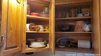 Two kitchen cabinets open containing a variety of plates, glass bowls, plastic food containers, wooden and plastic spoons, and other kitchen items.