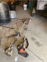 Two gas powered weed wackers lying on a concrete floor, showing overall used condition with some dirt and wear.