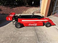 Full side view of red go-kart showing Coca-Cola side logo, body shape, wheels and engine at rear.