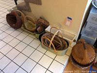 Photo showing a variety of baskets lined up against the wall on tile and wood floor including a round covered basket, a green metal container holding smaller baskets, and a wooden oblong box.