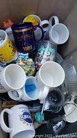 Photo of various coffee mugs showing different branded and printed designs including one with 1989 date, some white ceramic mugs, plastic cup with cartoon design, and a clear glass bowl.
