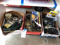 Photo showing three boxes filled with assorted vintage technology including modems, routers, power cables, phones, and electrical components.