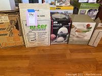 Four boxed kitchen appliances: Osterizer blender, Health Care juicer, Tropical Ice Shaver, and ovenware set arranged on floor.