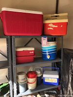 Photo of metal shelving unit holding an assortment of various coolers and beverage containers in red, blue, white and beige colors, showing condition and quantity.