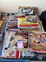 Photo showing multiple vintage board games stacked, including Operation, Monopoly, Chinese Checkers, Chutes and Ladders, Booby Trap, Balloon Sticker Fun, Kensington, and calligraphy and craft supplies along with packaging and pieces visible.