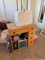 Sewing table with white plastic sewing machine case on top, storage space filled with various items below the table.