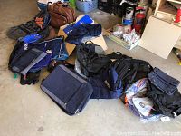 View of assorted sport and travel bags mostly in dark colors arranged on floor with some visible packing materials and boxes in background