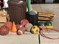 Photo showing various balls (basketballs, footballs, soccer balls), orange cones, helmets, and skateboards arranged on the ground near trash cans.